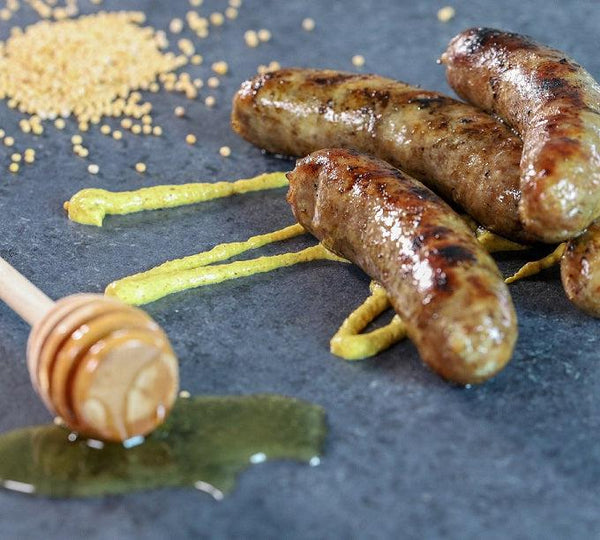 Honey Mustard Brats piled on one another with a mustard drizzle on them. To the left is a honey stick with honey dripping on the table, and Honey Mustard seasoning in the back left hand corner.